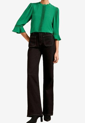 Femme portant un chemisier vert à manches froufroutantes et un pantalon large taille haute noir, assorti à des bottines à talons noires.