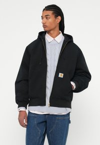 Mand med fletninger iført en sort zip-up hoodie over en lys blå kabelstrikket sweater og blå jeans, der kigger til venstre.