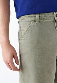 Light olive green trousers with a textured finish, featuring angled pockets, a button closure, and a fitted waistband.