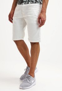 Shorts en denim blanc avec une coupe droite, un ourlet retroussé et des poches, associés à des chaussures de sport grises arborant un logo blanc.