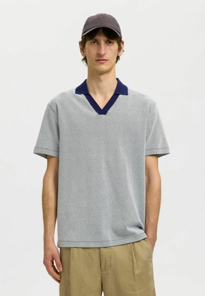 Homme portant une chemise grise à manches courtes avec col foncé, pantalon beige et casquette grise, debout avec une main dans la poche sur fond blanc.