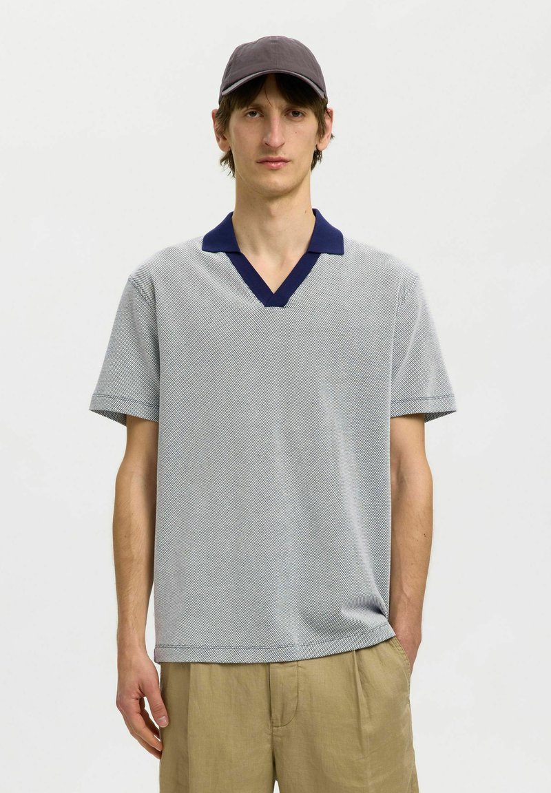 Homme portant une chemise grise à manches courtes avec col foncé, pantalon beige et casquette grise, debout avec une main dans la poche sur fond blanc.