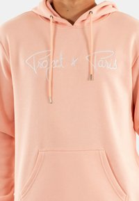 Hoodie rosa chiaro in tessuto morbido, dotato di tasca frontale e coulisse. Testo ricamato "Project x Paris" in bianco.