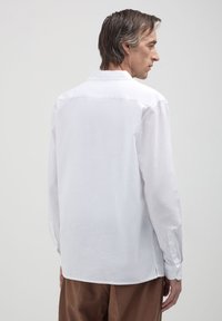 Uomo in piedi di spalle, che indossa una semplice camicia bianca a maniche lunghe e pantaloni marroni, su uno sfondo grigio uniforme.
