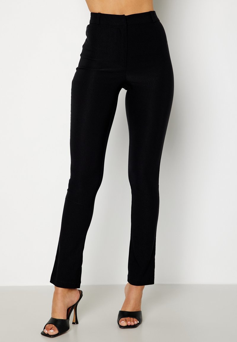 Pantalon noir slim porté avec des sandales à talons hauts noires ouvertes sur un fond uni.