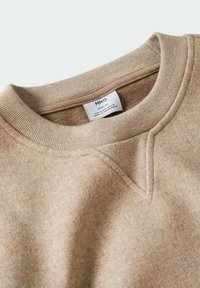 Beżowa bluza z okrągłym dekoltem, ściągaczami na rękawach i detalem w kształcie litery V przy kołnierzu. Miękka faktura z metką widoczną wewnątrz.