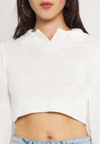 Vitt kort tröja med ribbat mönster, med V-ringning och platt krage. Designen inkluderar långa ärmar och en åtsittande midja.