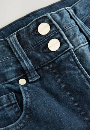 Jeans din denim de culoare bleumarin închis, cu un design cu talie înaltă, două butoane metalice argintii și detalii cu cusături în contrast. Buzunar frontal vizibil.
