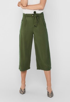 Kobieta ubrana w oliwkowozielone szerokie spodnie o długości do połowy łydki z paskiem przewiązanym w kokardę, do tego nosi buty na niskim obcasie wzorzyste na wzór skóry węża oraz biały top.