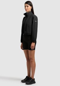 Ženska stoji bočno, oblečena v črno kratko jakno, črne kratke hlače in črne superge na enobarvnem sivem ozadju.