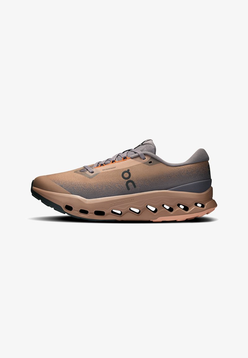 Chaussure de sport beige et grise avec logo noir, accents orange et semelle épaisse comportant plusieurs découpes ovales sur le côté.