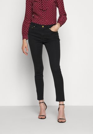 Frau trägt schwarze Slim-Fit-Jeans, rote gemusterte Langarmbluse und schwarze offene Pumps mit Knöchelriemen, steht mit einer Hand in der Hosentasche.
