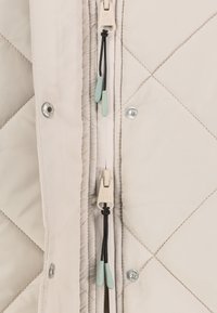 Gros plan sur le devant d'une veste matelassée beige avec une double fermeture éclair comportant des tirettes en tissu et des boutons-pression métalliques sur la patte de boutonnage.