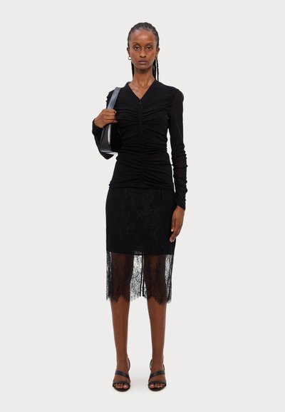 Top negro de manga larga con detalle fruncido, combinado con una falda lápiz de encaje negra que presenta un dobladillo transparente. La modelo sostiene un bolso negro y lleva tacones con tiras.