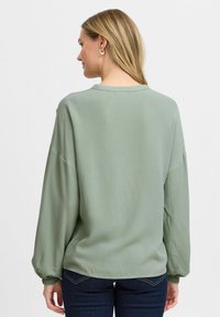 Maglione verde a maniche lunghe con una vestibilità ampia, scollo rotondo e polsini arricciati, abbinato a jeans in denim scuro.