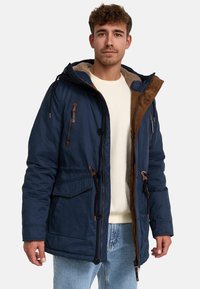 Navy blue parka jacket with a brown corduroy collar, zip and button closures, multiple pockets, and a soft, fleece-lined interior.