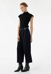 Musta hihaton paita korkealla kauluksella, yhdistettynä leveälahkeisiin mustiin housuihin ja teräväkärkisiin mustiin nilkkabootseihin, joissa on teksturoitu vyö.