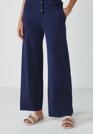Femme portant un pantalon large bleu marine avec des fentes latérales, des sandales beige à enfiler, et un haut assorti bleu marine à boutons.