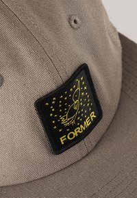 Grå keps med en svart rektangulär lapp med gul brodyr. Lappen visar ordet "FORMER" och en stiliserad fågidesign bland stjärnor.