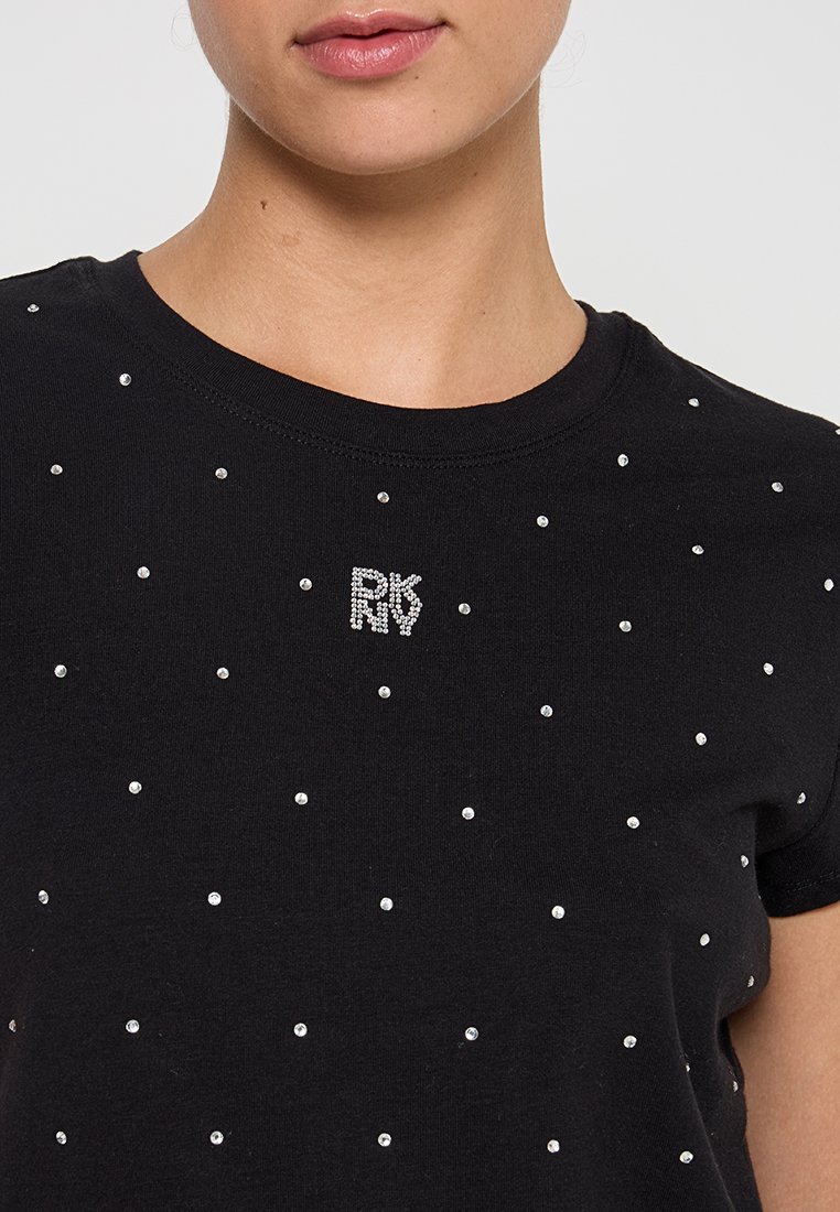 Femme portant un t-shirt noir avec de petites rivets en argent et un logo en strass "RDNY" centré sur la poitrine.