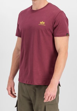 Mand iført en kortærmet vinrød t-shirt med gul Alpha Industries-logo og olivengrønne cargobukser.