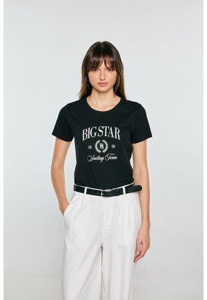 Kobieta w czarnej koszulce "BIG STAR Sailing Team" schowanej w białe spodnie z wysokim stanem i czarnym paskiem, stojąca na jednolitym tle.