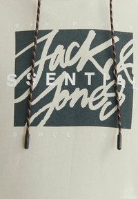 Lys beige hættetrøje med stort grafisk tryk i mørkegrøn og hvid, der viser teksten "Jack & Jones" og en justerbar snor med sorte og orange detaljer.