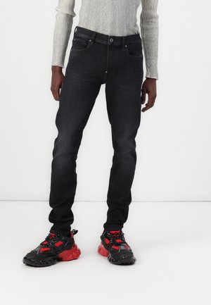 Personne portant un jean noir slim, des baskets épaisses rouges et noires, et un pull gris à côtes à manches longues sur un fond blanc uni.
