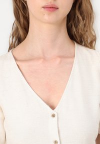 Femme aux cheveux châtain clair et ondulés portant un haut crème à encolure en V boutonné, vue du menton à la poitrine, sur un fond clair et uni.
