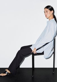 Light blue oversized button-up shirt, black wide-leg trousers, black slide sandals, smooth fabric, minimalist design, seated on a black chair.