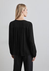 Zwarte blouse met lange mouwen, geplooide achterkant, iets afgeronde halslijn en een gestructureerde stof; gecombineerd met zwarte broek.