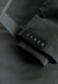 Schwarzer Jackenärmel mit einem Klettpatch mit der Aufschrift "BAKER", ausgestattet mit strukturiertem Stoff, einem eleganten Design und einer glatten Oberfläche.