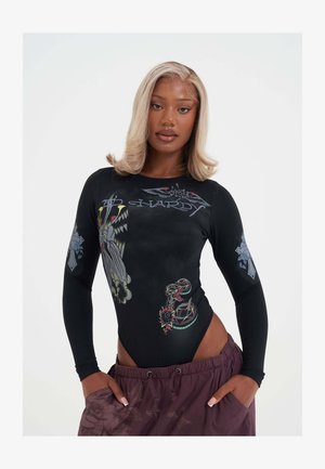 Jeune femme aux cheveux blonds mi-longs portant un body noir à manches longues avec des motifs style tatouage et un pantalon violet à motifs floraux.