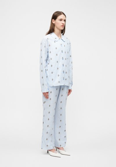 Woman in light blue striped pajama set with small bee patterns, standing with arms by sides, wearing white pointed heels on plain background.