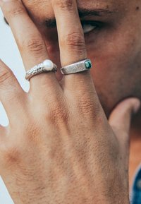 Två ringar på en hand: en silverring med en pärla, den andra silver med en strukturerad yta och en grön sten. Huden är synlig.