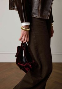 Burgundy velvet high-heeled shoes with a bow detail, held by a hand wearing a gold bangle, paired with dark brown pants and a brown leather jacket.