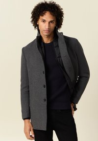 Cappotto di lana grigio con colletto alto, bottoni monopetto e fodera interna con zip. Indossato sopra un maglione a trama blu navy.