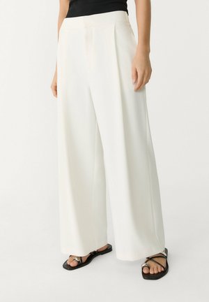 Frau trägt hochtaillierte, weit geschnittene weiße Hose, schwarzes ärmelloses Oberteil und schwarze flache Sandalen mit dünnen, sich kreuzenden Riemen und silbernem Ringdetail.