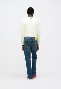 Άτομο που στέκεται με την πλάτη προς την κάμερα, φοράει λευκό πουλόβερ, μπλε τζιν με κίτρινη ετικέτα και κόκκινα αθλητικά παπούτσια, πάνω σε απλό λευκό φόντο.