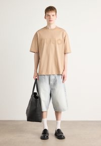 T-shirt bej oversized cu text embossat "HUGO", combinat cu pantaloni scurți din denim deschis. O geantă de umăr neagră și loafers negri completează ținuta.
