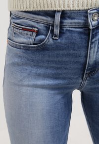 Närbild av ljusblå denimjeans i kombination med en krämfärgad stickad tröja, som visar detaljerna på framlappen och knappen.