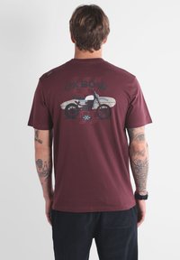 Homme avec des tatouages portant un t-shirt bordeaux avec un graphique de moto et de planche de surf, portant le texte "OXBOW" dans le dos.