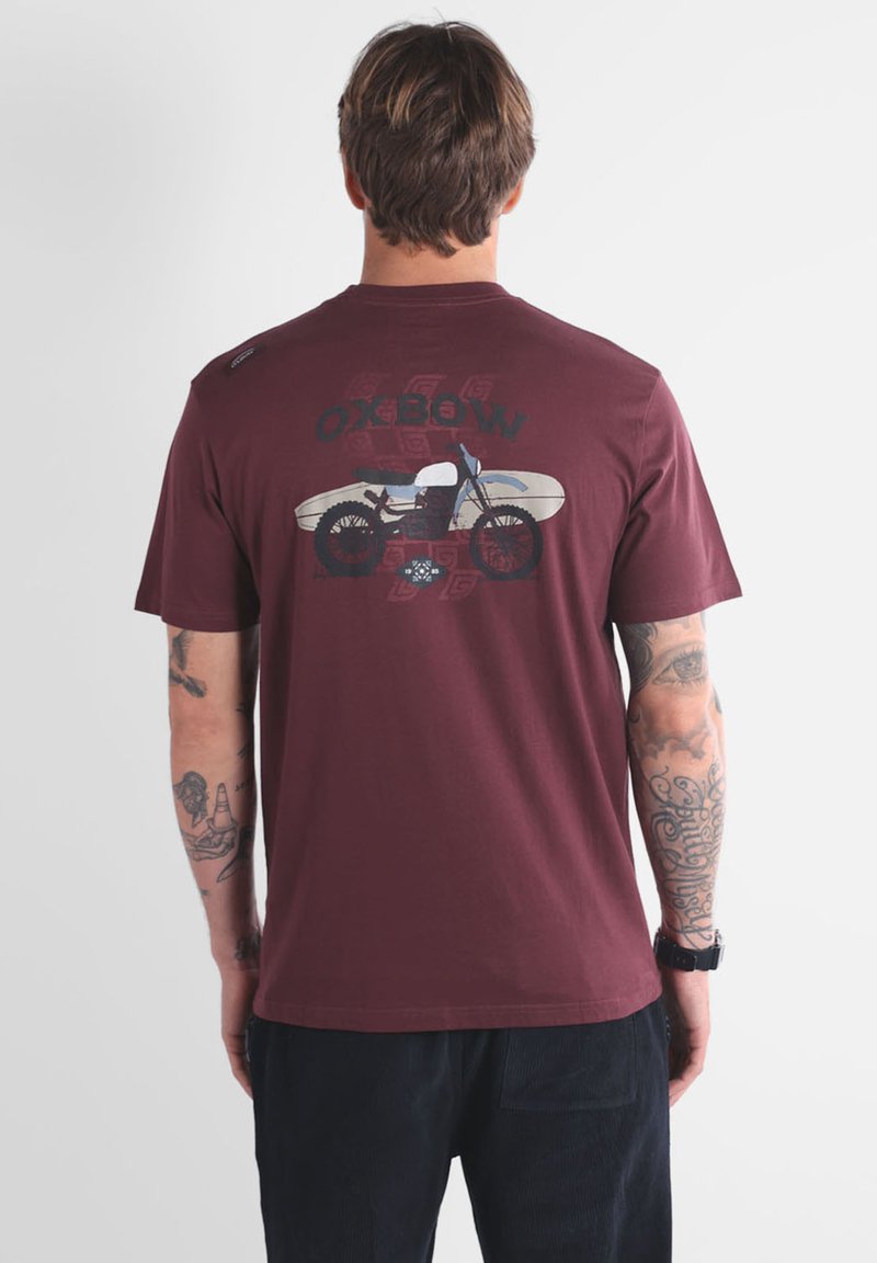 Homme avec des tatouages portant un t-shirt bordeaux avec un graphique de moto et de planche de surf, portant le texte "OXBOW" dans le dos.