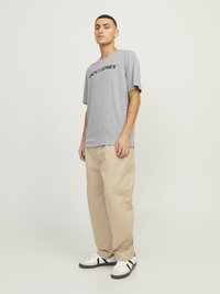 Grå t-skjorte med korte ermer som har en svart logo, sammen med lyse beige løstsittende bukser og hvite joggesko med svarte detaljer.