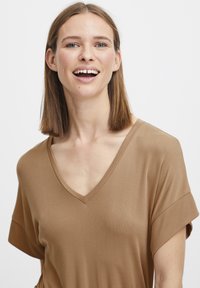 Hnědý top s krátkým rukávem ve tvaru V vyrobený z hladkého materiálu s volnějším střihem a jemnou strukturou rib. Modelka se usmívá a ukazuje přirozený make-up.