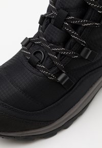 Zwarte wandelboot met textiel en rubberen zool. Bevat dikke zwarte veters, zwarte plastic details en reflecterende witte accenten.