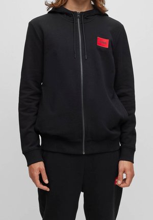 Person iført sort hættetrøje med lynlås og matchende sorte bukser med røde rektangulære logo patches på bryst og lår.