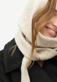 Fular din lână bej răsucit în jurul gâtului, cu o textură moale. Purtați cu un pardesiu negru, oferind un aspect natural și discret.