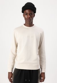 Beige genser laget av et mykt materiale med lange ermer, rutete krage og mansjetter; har liten, subtil brodert tekst på fronten.
