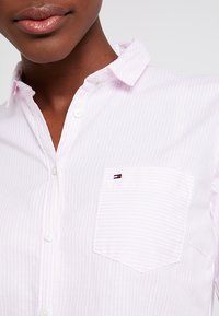 Camicia button-up rosa chiaro con righe bianche, colletto e tasca sul petto con un piccolo logo. Tessuto dal tocco morbido.